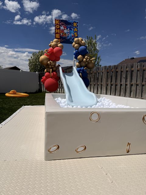 Superhero-themed slide and ball pit with red, blue, and gold balloons